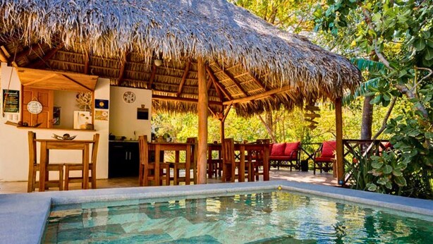 A pool area with a thatched-roof structure, wooden furniture, and a clear pool. - Antema Lodge (Tamarindo)