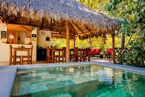A pool area with a thatched-roof structure, wooden furniture, and a clear pool. - Antema Lodge (Tamarindo)