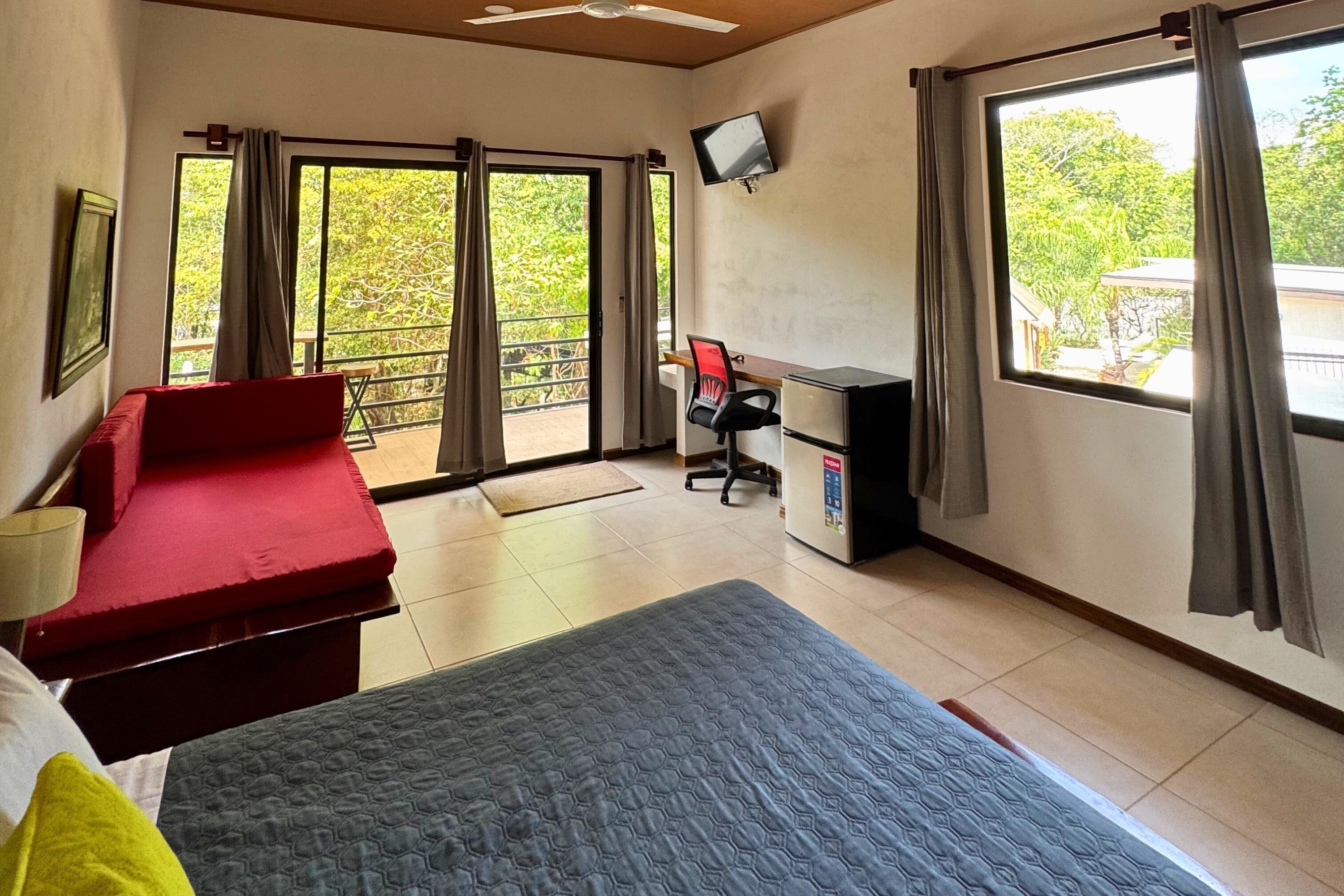 Una stanza con un letto, una scrivania, una sedia e una televisione. Sono presenti ampie finestre con tende, un balcone e vista sul verde.