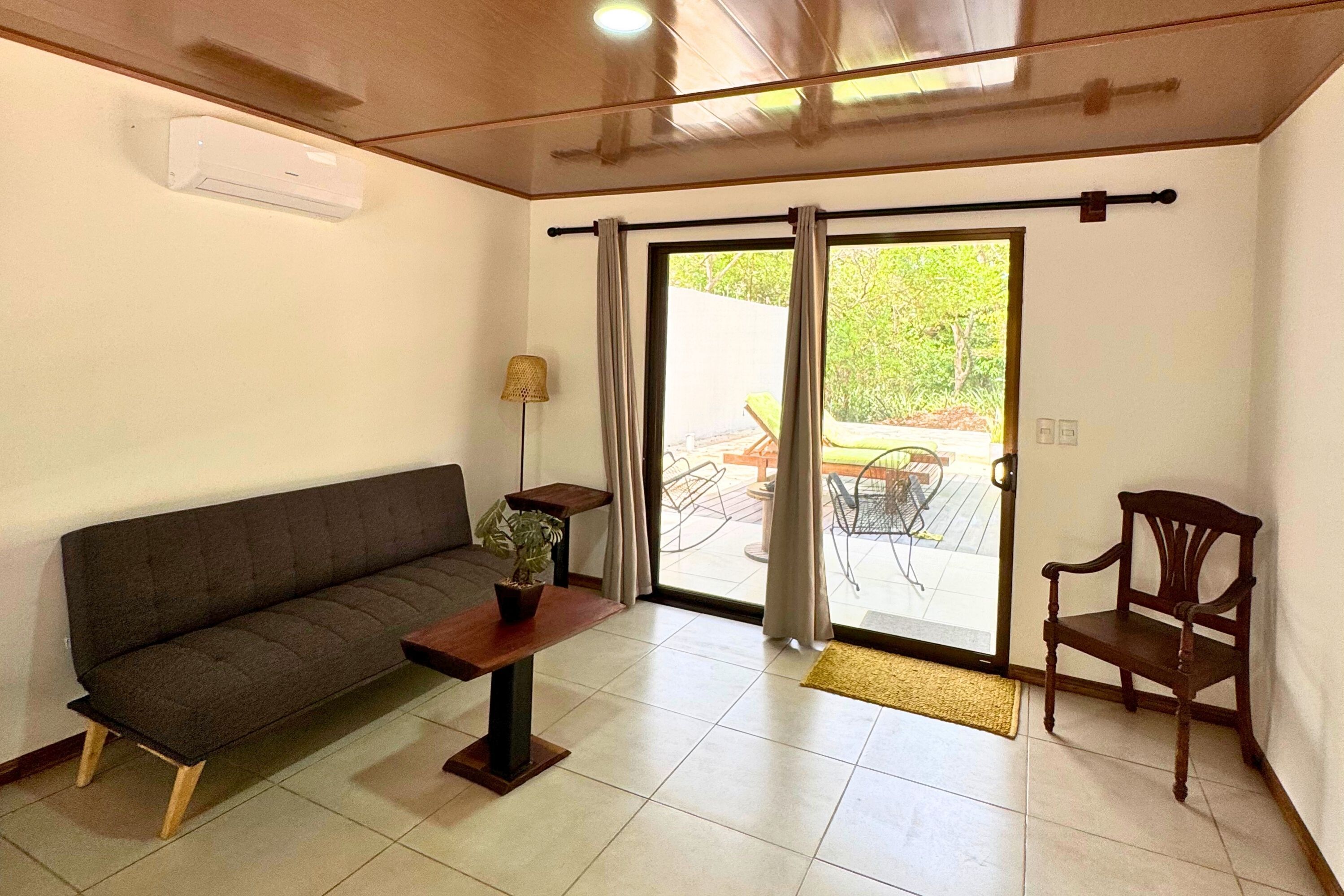 A living room with a sofa, a small table with a plant, a chair, and a sliding glass door leading to an outdoor area.