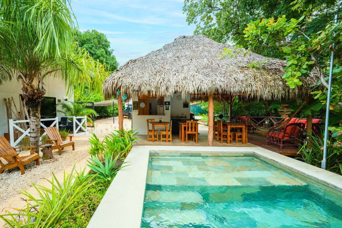 A pool area with a thatched-roof structure, wooden chairs, and a dining table.