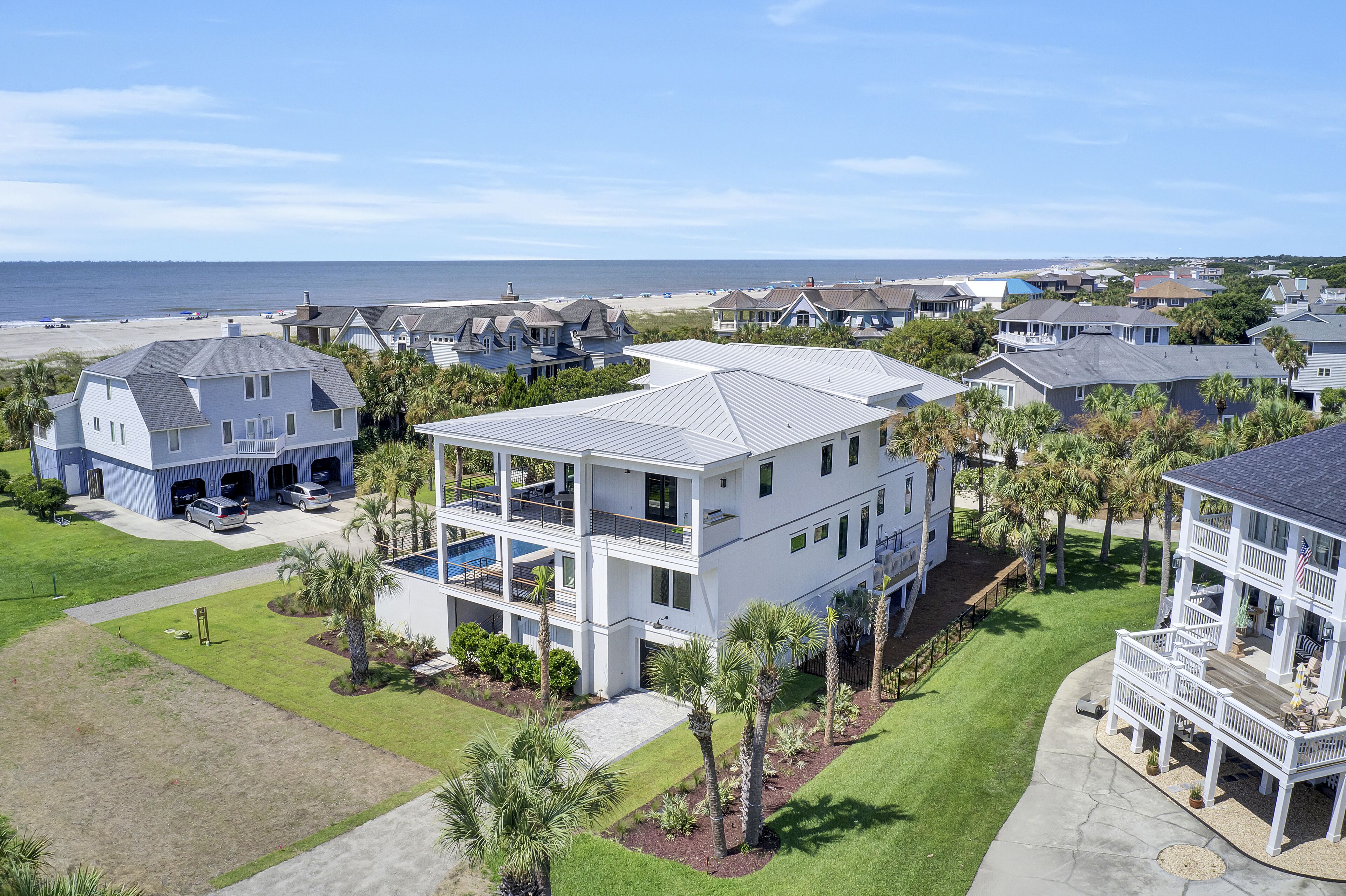 A modern two-story house with a large balcony, surrounded by palm trees and a view of the beach.