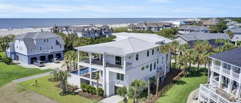 A modern two-story house with a large balcony, surrounded by palm trees and a view of the beach.