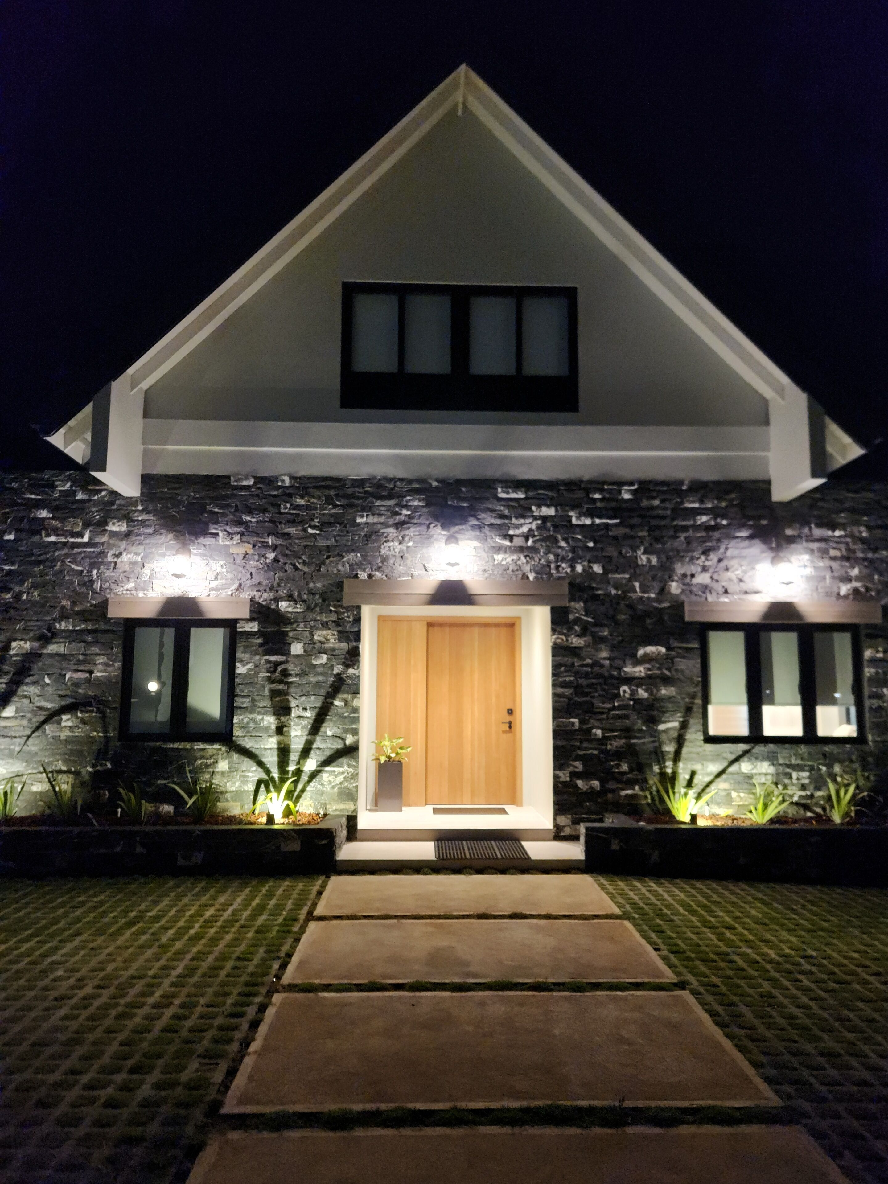 A modern house with a stone facade, a wooden front door, and two lit windows.