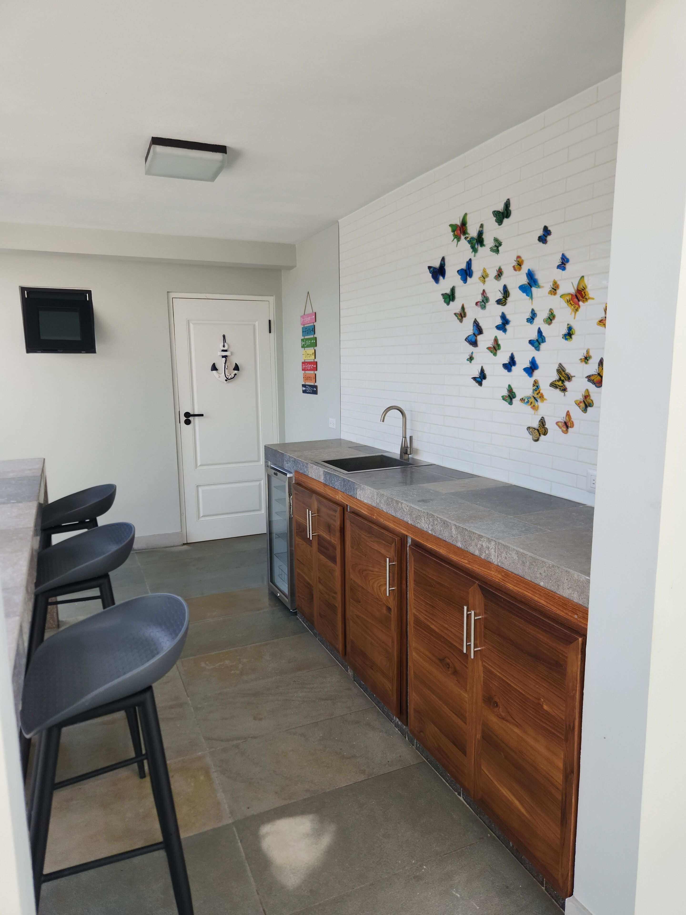 Een keuken met een houten aanrecht, een spoelbak en een spatwand met vlinderstickers. Drie barkrukken en een deur met slot.