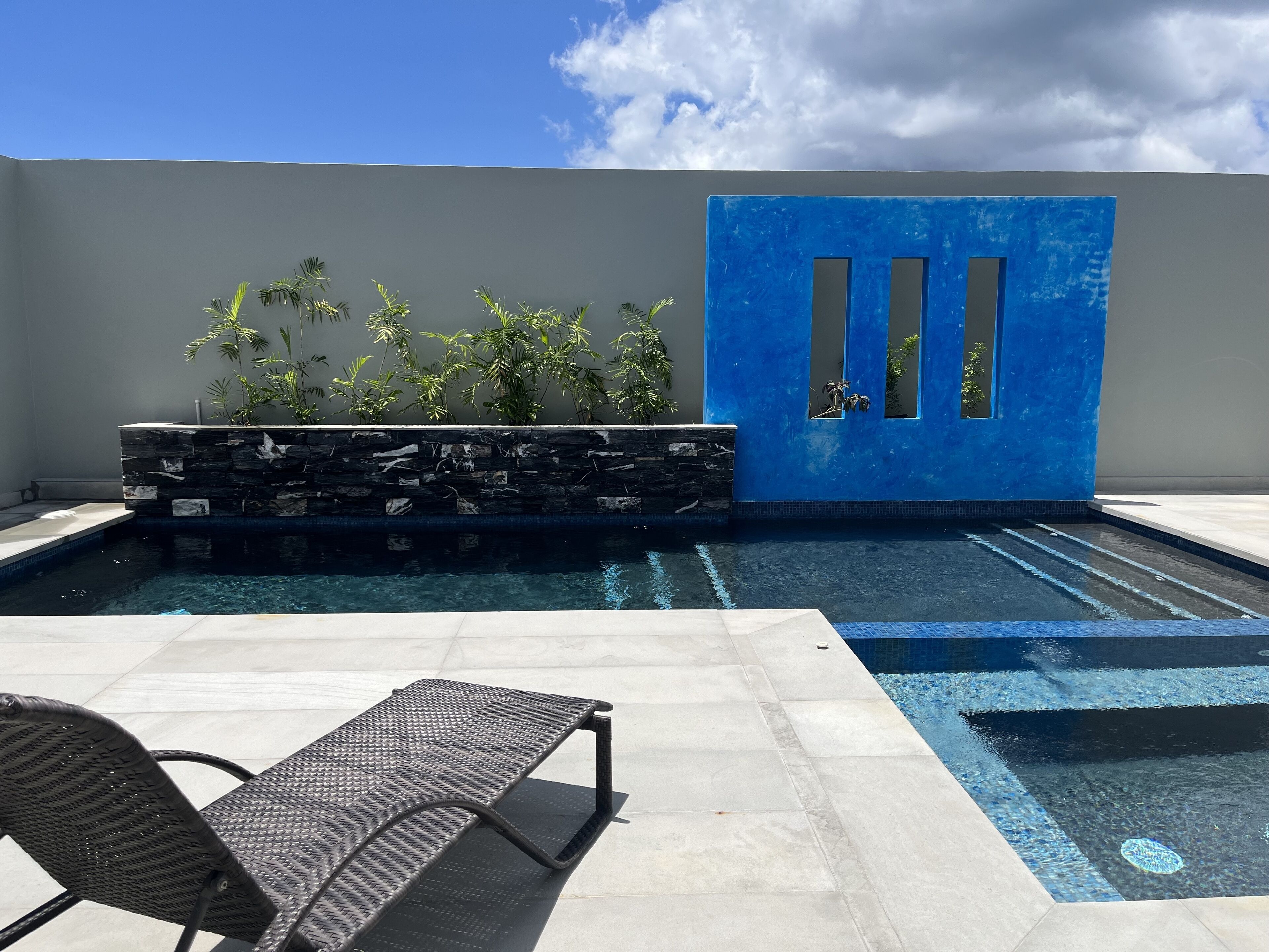 A swimming pool with a stone wall and plants, a blue door, and a lounge chair.