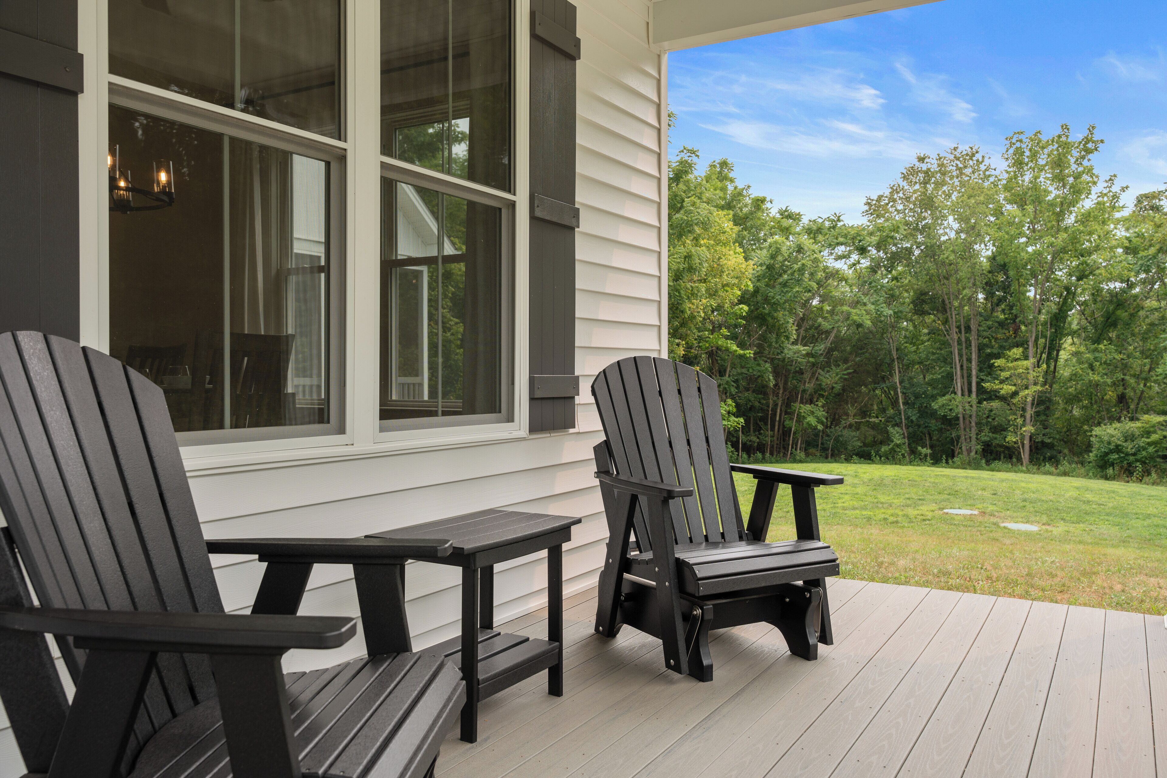 A deck with two Adirondack chairs and a small table, overlooking a grassy yard and trees.