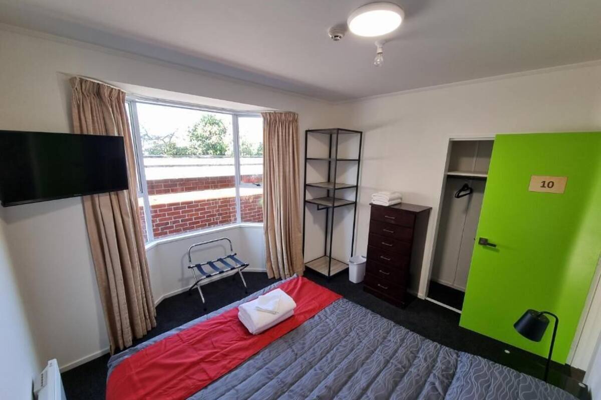 A hotel room with a bed, a TV, a window with curtains, a green door, and a small shelf.
