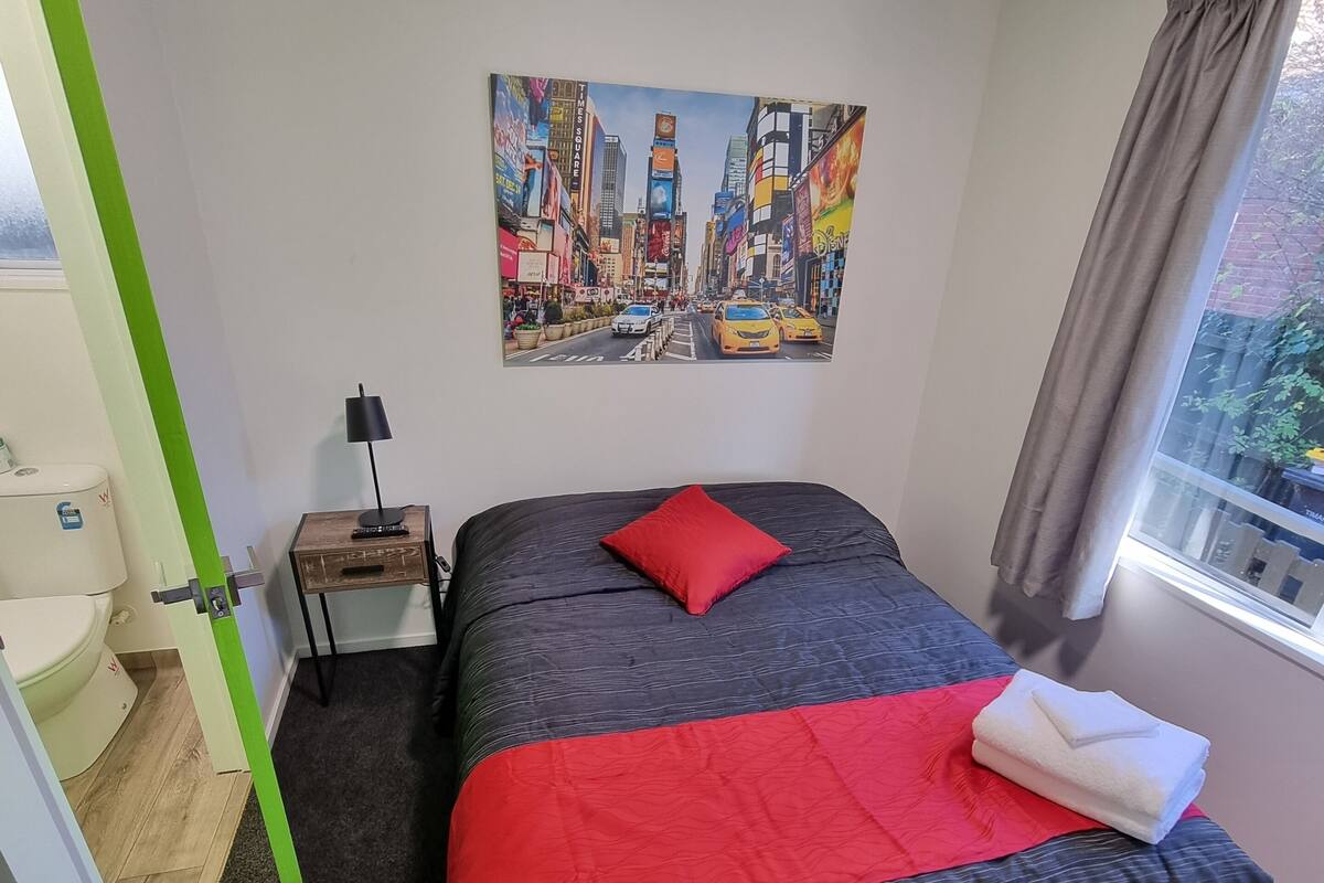 A bedroom with a bed, a painting of a cityscape, a nightstand, a lamp, and a window with curtains.