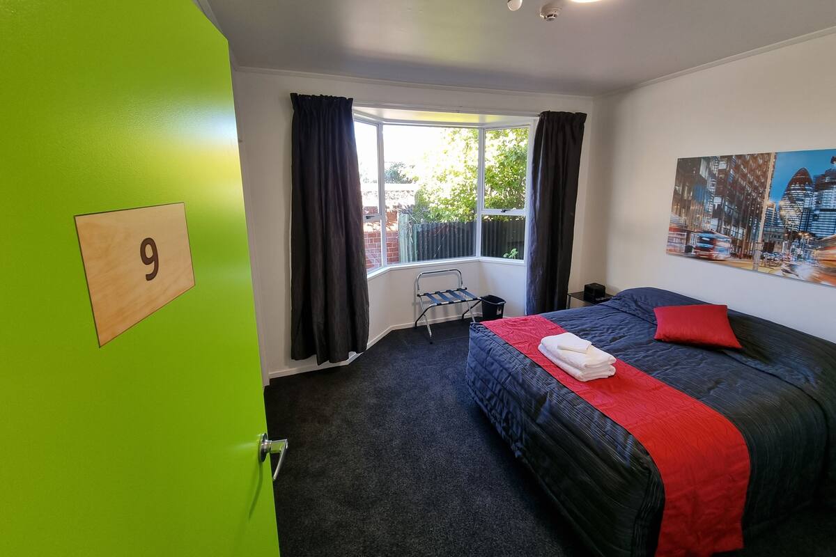 A hotel room with a large bed, a window with curtains, a radiator, and a wall-mounted number sign.