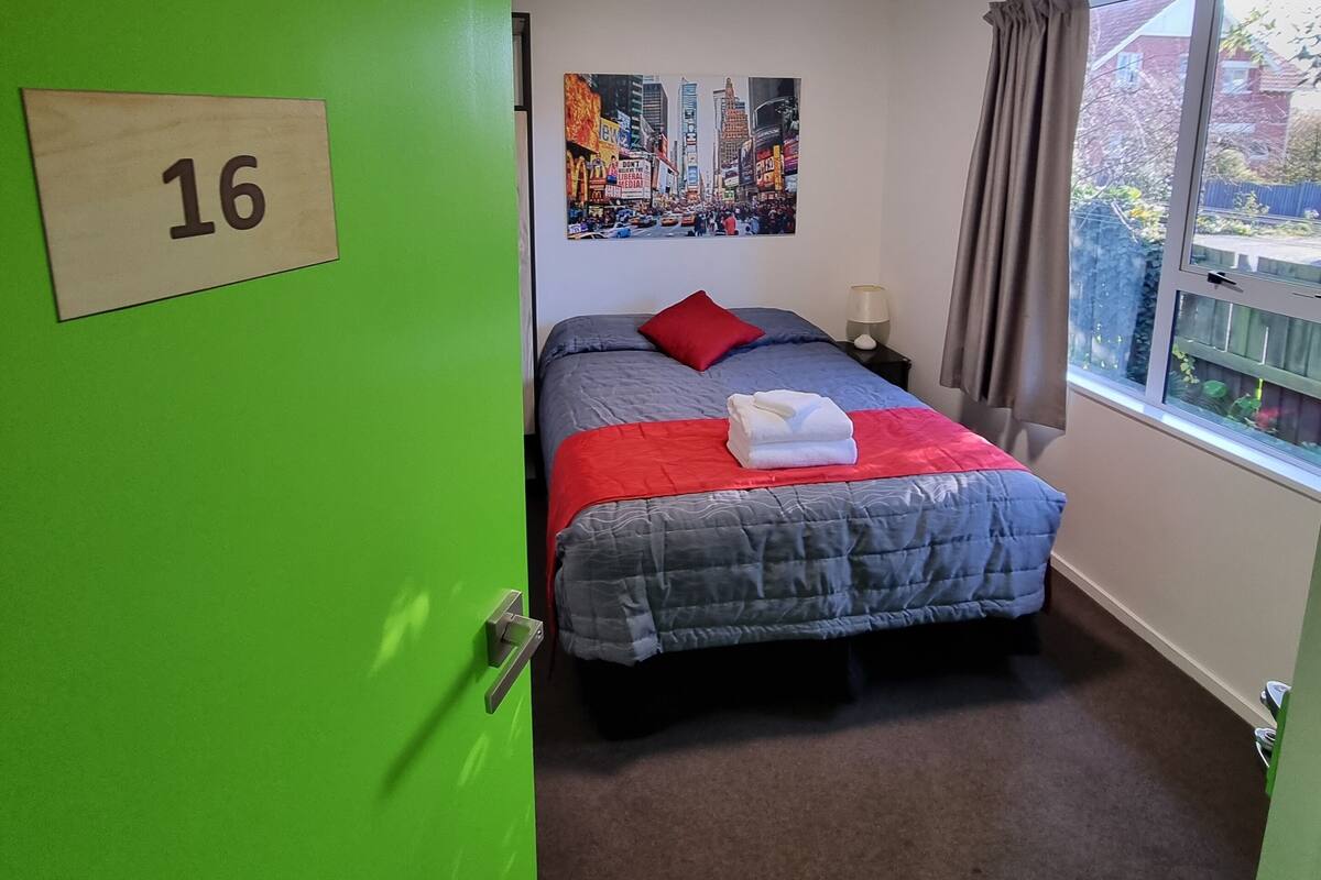 A room with a green door marked '16', a bed with a red pillow, a blue blanket, and a red throw, a window with curtains, and a poster of a cityscape on the wall.