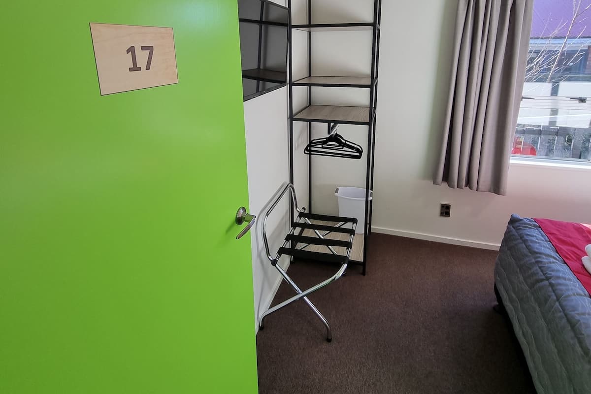 A room with a green door marked with the number 17, a metal rack with hangers, a bed with a red blanket, and a window with curtains.
