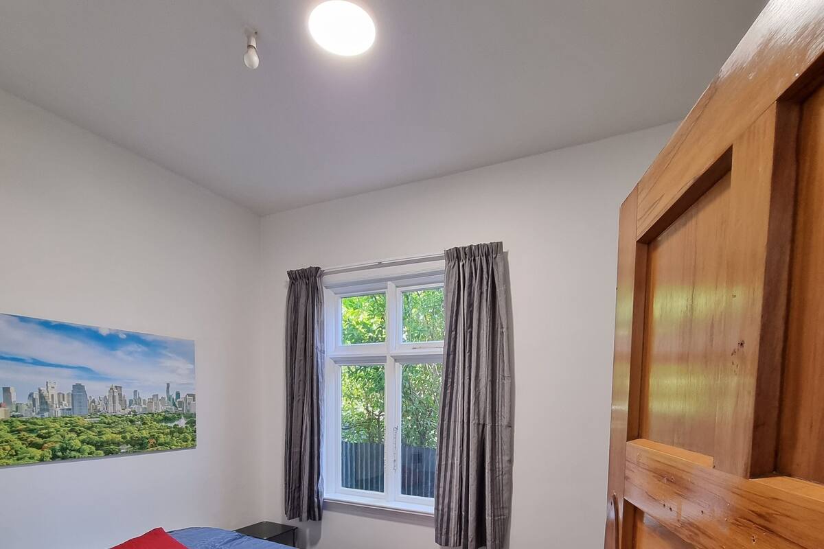A bedroom with a bed, a window with curtains, a painting on the wall, and a wooden door.