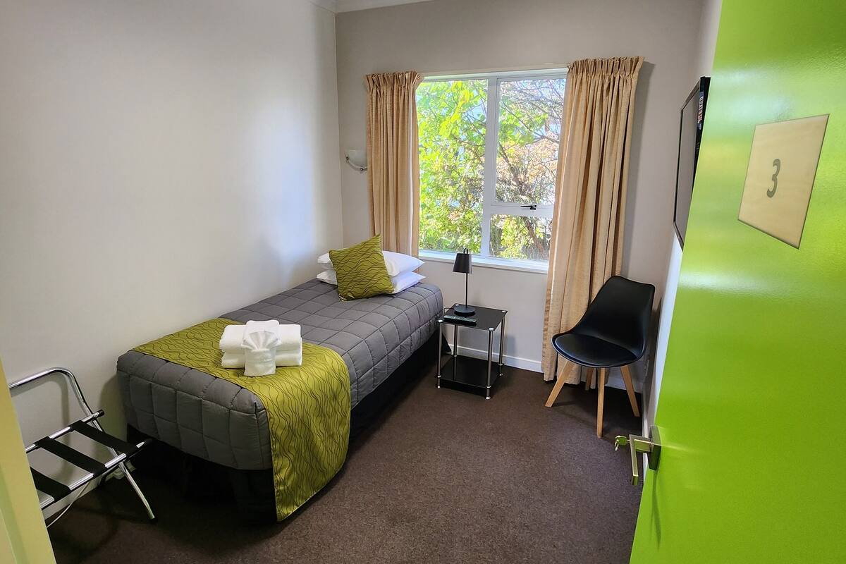 A compact room with a single bed, a small table, a chair, and a window with curtains.