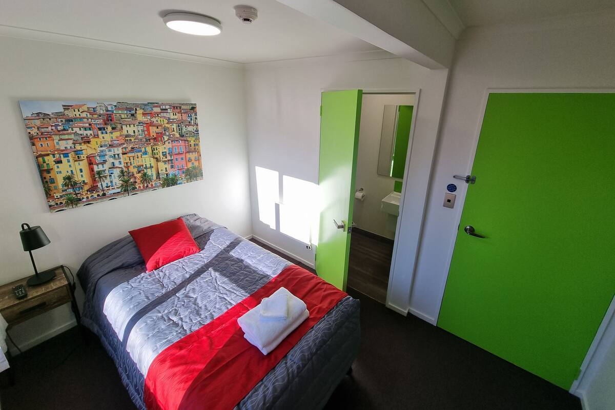A hotel room with a bed, a nightstand, a painting, a green door, and a bathroom visible through an open door.