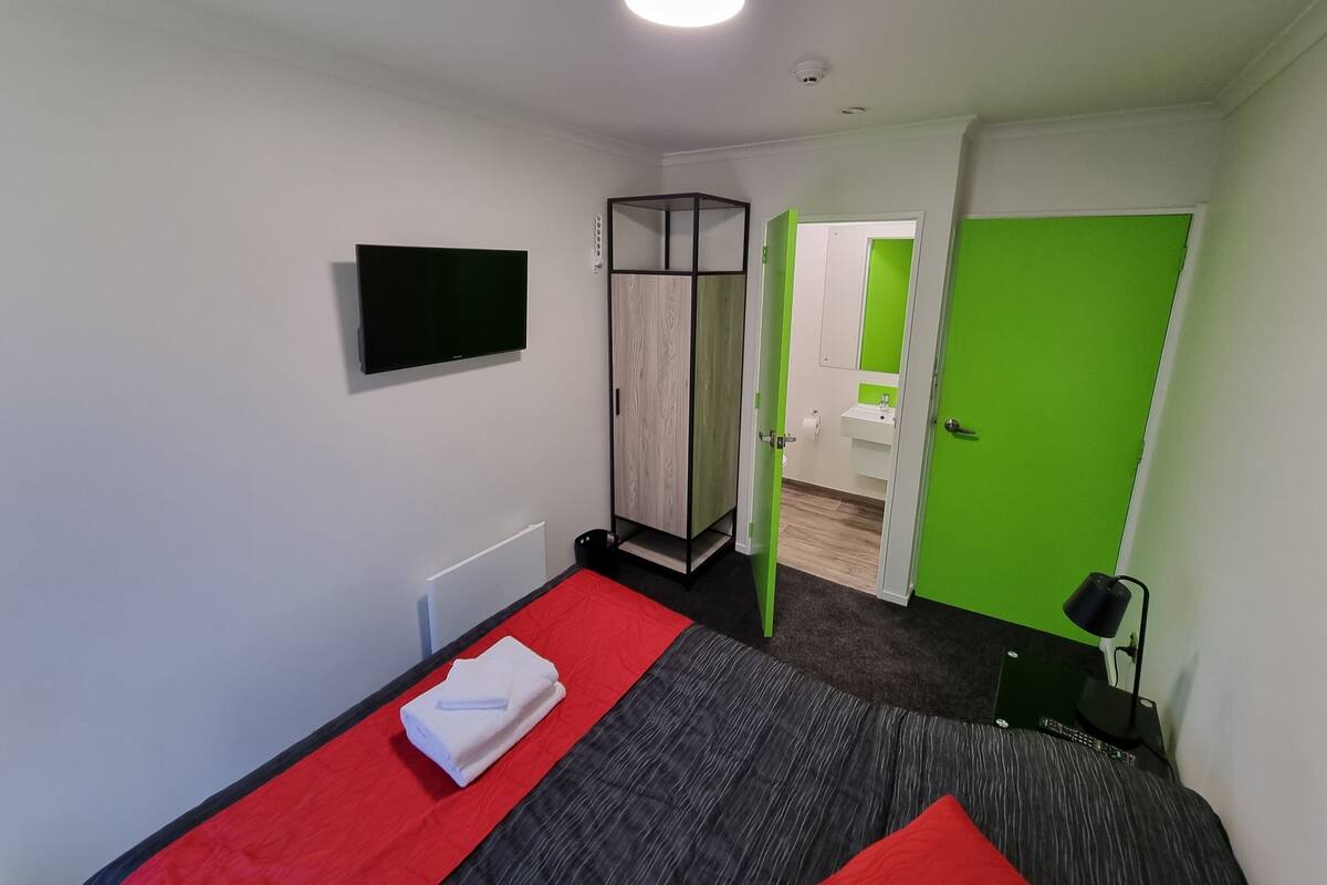 A modern hotel room with a bed, a TV, a wardrobe, and a bathroom visible through an open door.