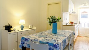 Un pequeño comedor con una mesa dispuesta para dos personas, un jarrón azul con flores, una lámpara y una cocina al fondo.