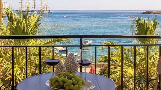 Un balcón con una mesa preparada para dos personas, con vista a la costa y barcos.