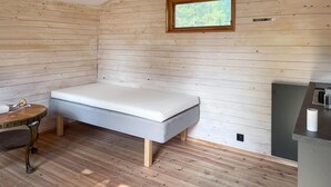 Una habitación pequeña con cama individual, paredes y suelo de madera, una mesita con lámpara y una ventana con vistas a árboles.