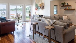 Un salón espacioso con techos altos, un ventanal grande con vistas a un lago, un rincón de estar acogedor con un sofá y sillones, y una escalera que conduce a un nivel superior.