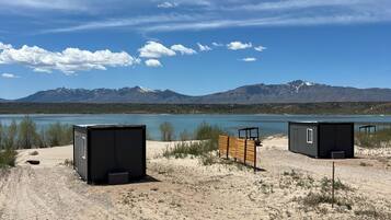 A desert landscape with two small, dark-colored cabins near a body of water, with mountains in the background.