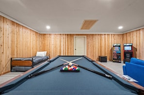 Une table de billard avec des billes, un canapé et deux bornes d’arcade dans une pièce aux murs en bois et dotée d’une porte.