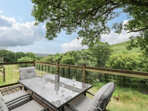 An outdoor patio with a glass-top table and chairs, surrounded by a wooden deck and a view of a green landscape. - Nant Glas (Llanwrda)