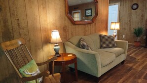 A cozy living room with wooden paneling, a green sofa with patterned cushions, a wooden rocking chair, and two table lamps. - Cozy Bear Rock Cottage with hot tub  (Monroe)