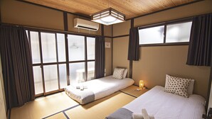 Ein traditionell japanisch gestalteter Raum mit Tatami-Böden, zwei Betten mit Kissen, einem Nachttisch mit Lampe und einem großen Fenster mit Vorhängen.