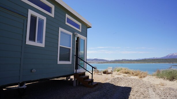 Uma casa móvel azul com porta da frente, um pequeno lance de degraus e vista para um lago e montanhas ao fundo.