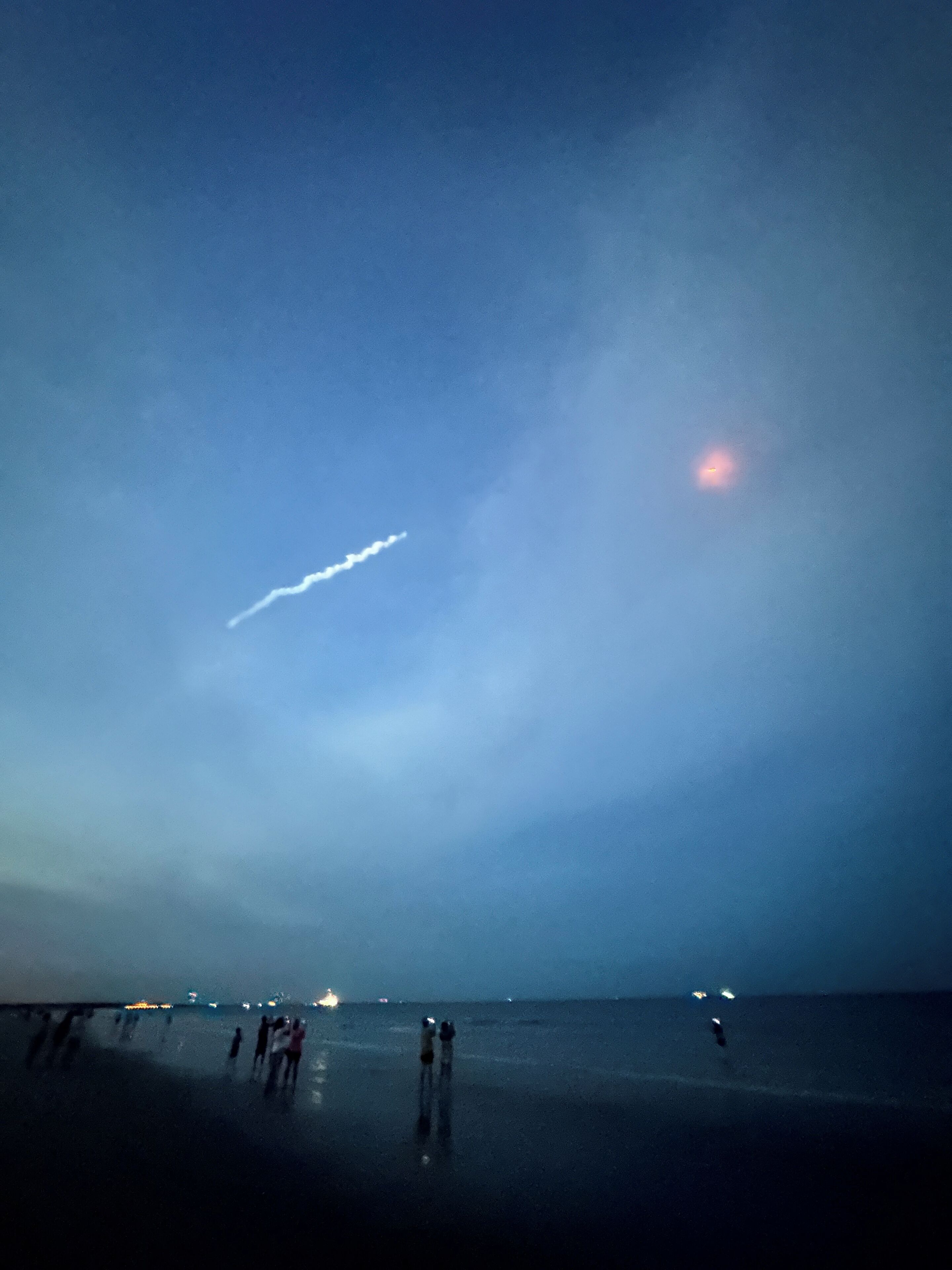 A beach at dusk with a visible contrail in the sky and a few people walking along the shore.
