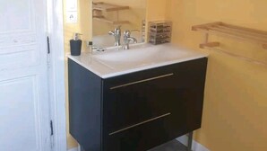 Un baño con una puerta blanca, un tocador negro con espejo, un lavabo blanco y una estantería de madera.