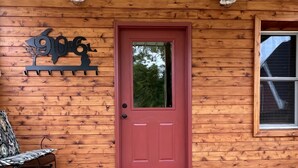 A wooden cabin with a red door, a bench, and a decorative sign. -  Lakeside Hideaway: Full-Home Retreat on the Remote Boney Falls Basin (Cornell)