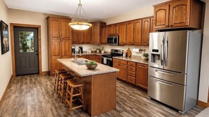 A kitchen with wooden cabinets, a central island with stools, stainless steel appliances, and a hanging light fixture. -  Lakeside Hideaway: Full-Home Retreat on the Remote Boney Falls Basin (Cornell)