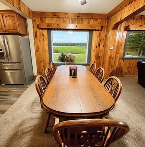 A wooden-paneled dining room with a large table and chairs, a refrigerator, and a view of a green landscape through a window. - Lakeside Hideaway: Full-Home Retreat on the Remote Boney Falls Basin (Cornell)