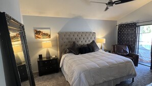 Une chambre à coucher avec une tête de lit capitonnée, un lit avec une literie blanche, un ventilateur de plafond, une table de chevet avec une lampe, une chaise et une fenêtre avec des rideaux.