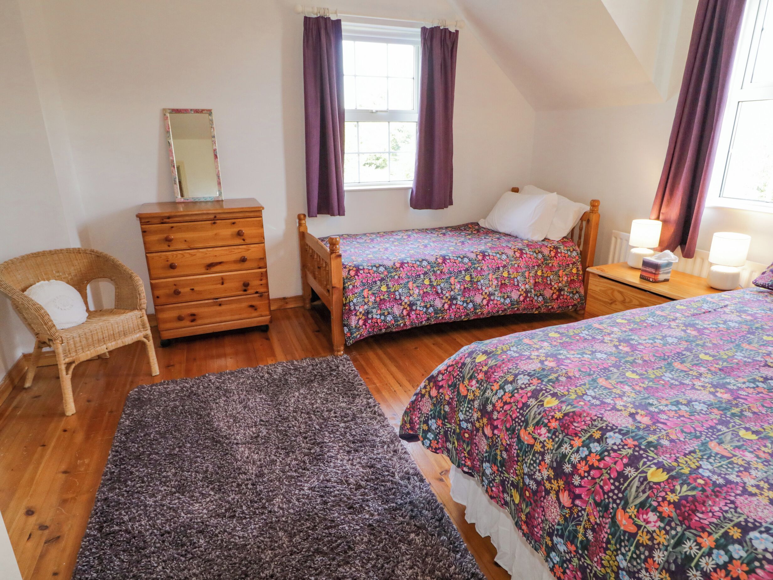 A bedroom with two single beds, a wooden dresser, a wicker chair, a window with purple curtains, and a nightstand with a lamp.