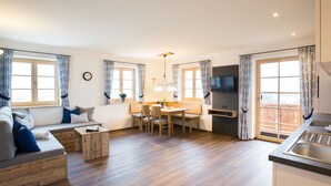 Ein modernes Wohnzimmer mit einer Couch, einem Esstisch und Stühlen. An der Wand ist ein Fernseher angebracht und es gibt ein Fenster mit Vorhängen.