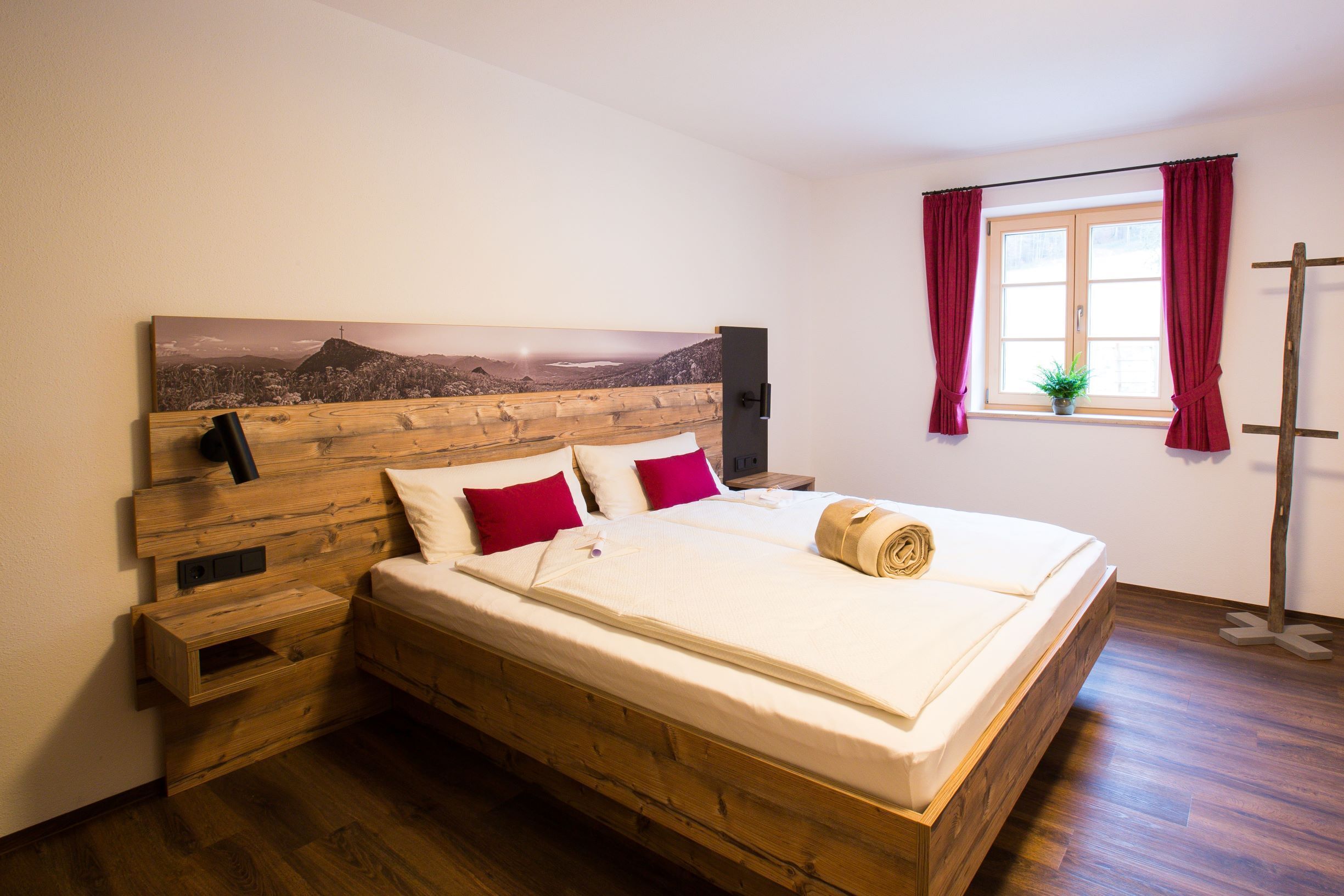 A wooden bed with a headboard, red pillows, and a wooden nightstand with a plant.