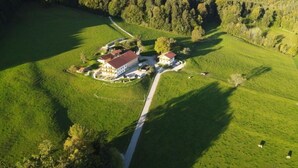 An aerial view of a rural property with a main building surrounded by green fields and a forested hill. - Almwiese für 2-4 Personen (Bernau am Chiemsee)