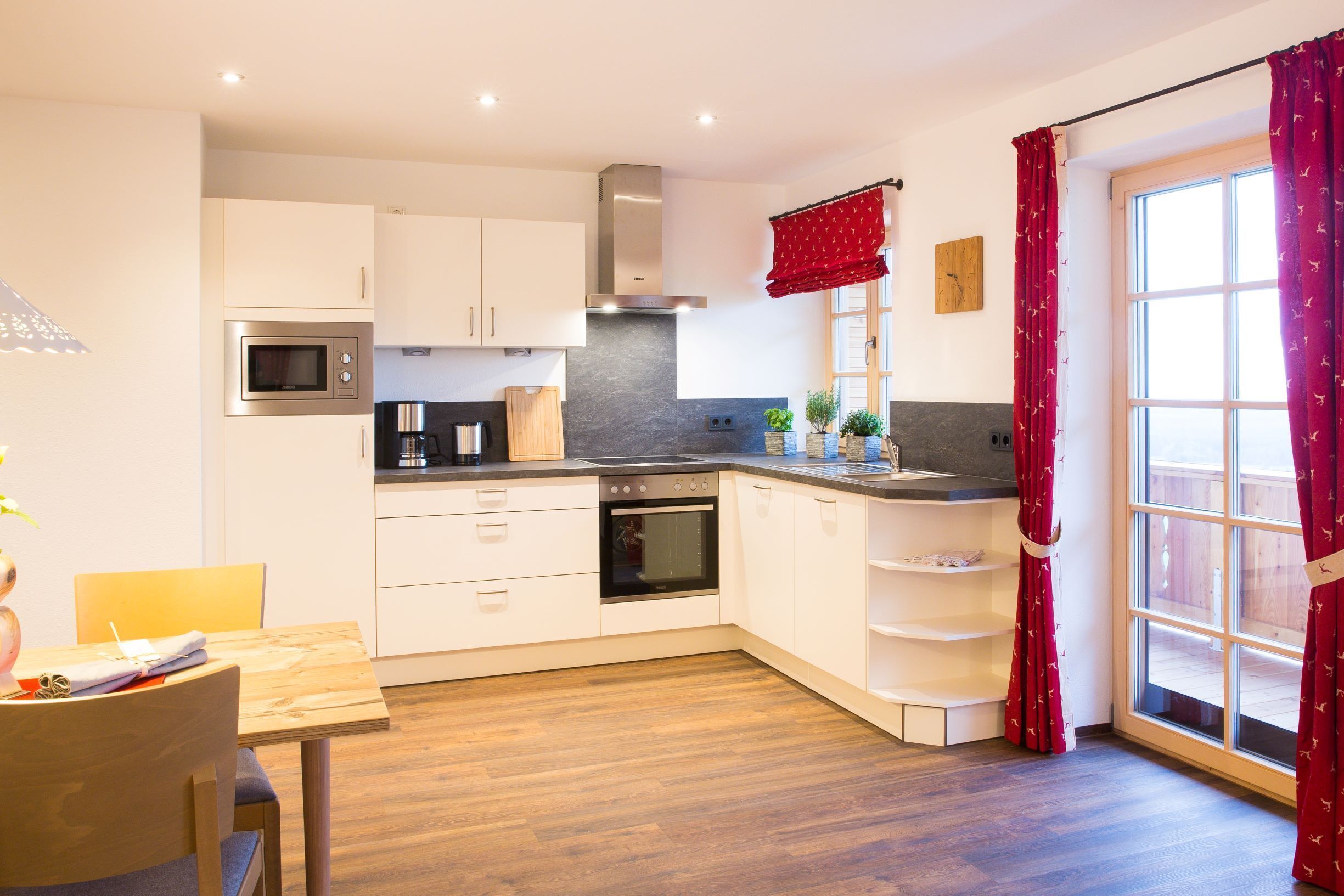 Eine moderne Küche mit weißen Schränken, einem Edelstahlbackofen und einer Mikrowelle. Es gibt einen Holztisch mit Stühlen und ein Fenster mit roten Vorhängen.