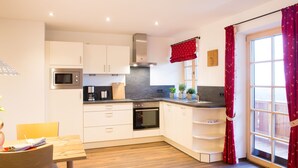 A modern kitchen with white cabinets, a stainless steel oven, and a microwave. There is a wooden dining table with chairs and a window with red curtains. - Almwiese für 2-4 Personen (Bernau am Chiemsee)
