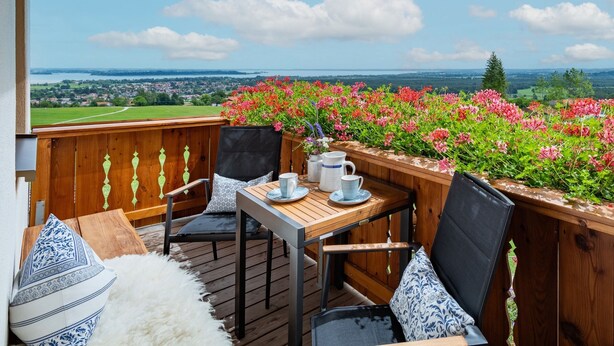 A wooden deck with a table and chairs, overlooking a scenic landscape with flowers and a distant view of houses and greenery. - Kräutergarten für 2-5 Personen (Bernau am Chiemsee)