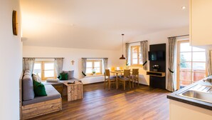 A modern living room with a wooden floor, a sofa, a dining table with chairs, a TV mounted on the wall, and large windows with curtains. - Kräutergarten für 2-5 Personen (Bernau am Chiemsee)