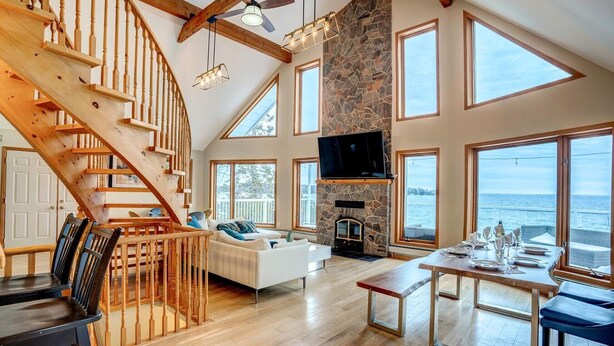 A spacious living area with a wooden staircase, a stone fireplace, and large windows overlooking a body of water. - Lake Simcoe Waterfront Lake House (Hawkestone)