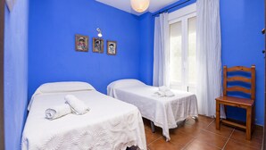Ein Zimmer mit zwei Betten, einem Holzstuhl, einem Fenster mit Vorhängen und Bildern an der Wand.