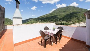 Eine Terrasse mit Tisch und Stühlen, eine weiße Wand und Blick auf einen grünen Hügel.