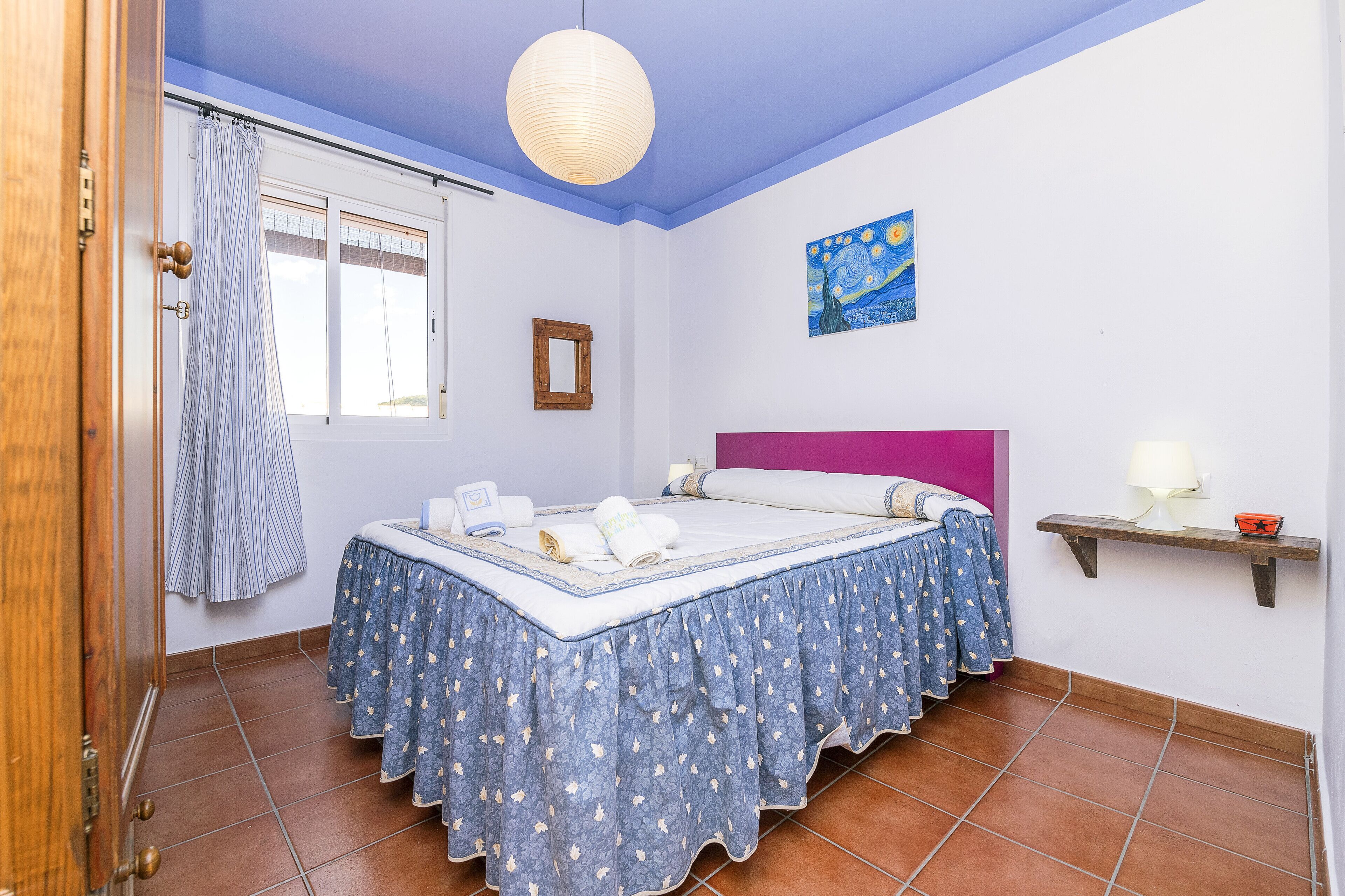 Une chambre à coucher avec un lit, une fenêtre avec des rideaux, un tableau au mur et une table de chevet avec une lampe.