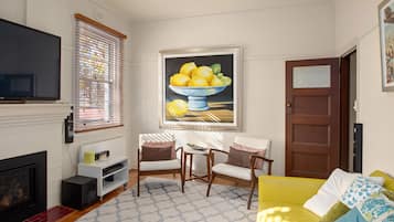 A living room with a fireplace, a television, a painting of lemons, two chairs, a yellow sofa, and a door.