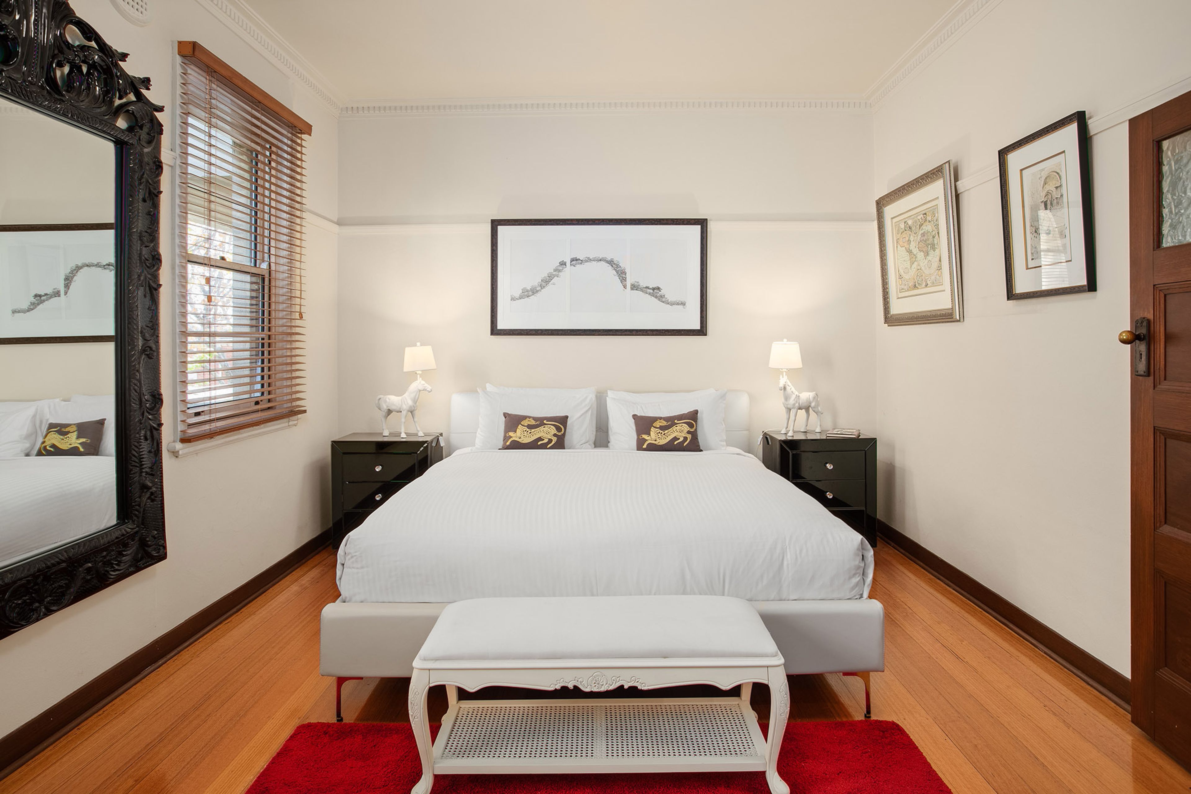 Habitación de hotel con una cama grande, dos mesitas de noche con lámparas, un banco, un espejo amplio y obras de arte en las paredes.